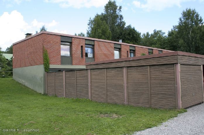 Two-story brick building behind a long row of brown wooden garages on a grassy lot. Trees and a blue sky in the background.